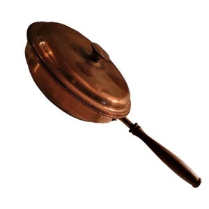 May include: A copper-coloured, oval-shaped pan with a lid and a long, dark wooden handle. The pan has a rounded lid with a small knob. The handle is attached to the pan with a metal piece. The pan is likely used for cooking.