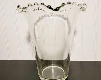 Könnte beinhalten: Eine klare Glasvase mit ausgestelltem Oberteil und dekorativem, perlenbesetztem Rand. Die Vase hat einen zylindrischen Körper und steht auf einer schwarzen Oberfläche vor einer weiß getäfelten Wand. Die Vase ist leer und scheint aus dickem Glas zu sein.