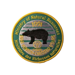 May include: Embroidered patch with a circular design. The patch features a black bear silhouette, the text "Ministry of Natural Resources" and "Bear Hunter" in green and yellow, and the year "1991". The patch has a green border.
