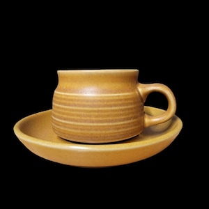 May include: A mustard-colored ceramic teacup and saucer set. The cup has a handle and horizontal ridged detailing. The saucer is round and shallow. The set is on a black background.