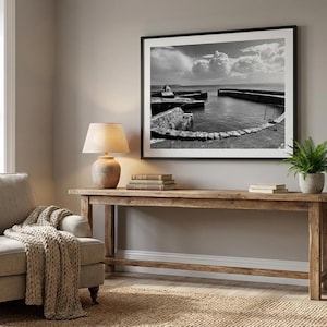 Puede incluir: Un salón con una fotografía en blanco y negro enmarcada de un puerto. Una consola de madera sostiene una lámpara, libros y un helecho en maceta. Un sillón con una manta de punto y una alfombra tejida completan el ambiente acogedor.