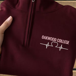 May include: A maroon quarter-zip sweatshirt with the text "OAKWOOD COLLEGE" and a heartbeat graphic in white. The sweatshirt has a silver zipper and is folded. The background is a wooden surface.