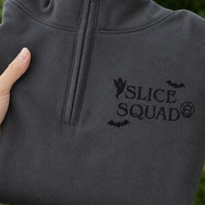 Peut inclure: Un sweat-shirt gris anthracite à fermeture éclair quart de col avec une broderie noire sur la poitrine. Le motif comprend le texte "SLICE SQUAD" entouré de petites icônes brodées représentant un fantôme, deux chauves-souris et une part de pizza.