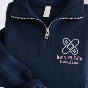 May include: A navy blue quarter-zip fleece sweatshirt with a white and pink graphic. The graphic includes a bandage design and the text "Jessica RN, CWCN Wound Care". The sweatshirt is folded on a light wood surface.