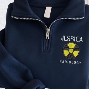 Puede incluir: Un suéter azul marino con cremallera corta que presenta un símbolo de radiación amarillo y negro bordado en el pecho. El texto JESSICA está bordado en blanco sobre el símbolo y la palabra RADIOLOGY está bordada en blanco debajo.