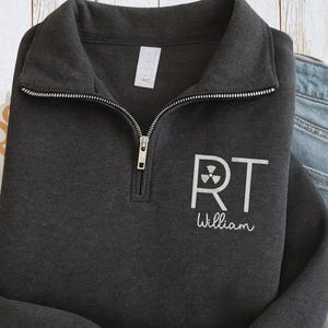 Può includere: Felpa grigio scuro con zip a un quarto di giro con un design bianco "RT William". Il design include un simbolo di radiazione. La felpa ha una cerniera argentata e un colletto ripiegato.