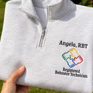 May include: A light gray quarter-zip sweatshirt featuring embroidered text that reads Angela, RBT and Registered Behavior Technician. Below the name is a colorful embroidered graphic of four hands in yellow, red, blue, and green joined together.