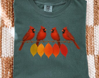 May include: A teal Comfort Colors t-shirt featuring a graphic of four red cardinals perched on a wire above colorful autumn leaves. The shirt is folded on a textured, neutral-toned surface.