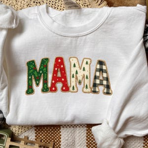 Könnte beinhalten: Weißes Sweatshirt mit dem Wort "MAMA" in dekorativen Buchstaben. Das "M" ist grün mit einem Blättermuster, das erste "A" ist rot mit weißen Sternen, das zweite "M" hat grüne Bäume und das letzte "A" ist schwarz-weiß kariert.