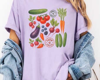 May include: A lilac t-shirt with a vibrant print of vegetables such as tomatoes, carrots, and cucumbers. The design incorporates the text "Farm Fresh" and "Locally Grown".