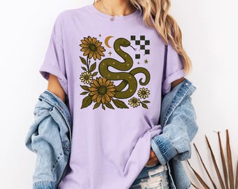 Könnte beinhalten: Helllila T-Shirt mit einem grünen Schlangen-Design, Sonnenblumen, Sternen und einem Mond. Die Grafik enthält ein kariertes Quadrat. Das Design ist auf einem weichen, bequemen T-Shirt gedruckt.