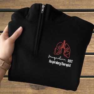 Puede incluir: Sudadera negra con cremallera de cuarto de cremallera, con un gráfico de pulmón rojo y el texto "Jacqueline, RRT Respiratory Therapist" en blanco. La sudadera tiene cremallera y está doblada.
