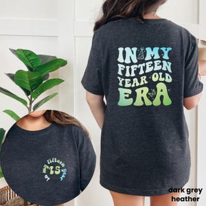 Peut inclure: T-shirt gris chiné foncé avec un design dégradé arc-en-ciel. Le dos du t-shirt affiche "In my fifteen year old era" avec un chapeau de fête et un papillon. Le devant du t-shirt a un cercle avec le nombre 15 en dégradé arc-en-ciel.