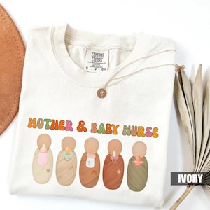 May include: Ivory t-shirt featuring the text "MOTHER & BABY NURSE" in retro orange and green lettering. Below the text are five cartoon babies wrapped in blankets, each with a different accessory. A gold necklace is draped across the shirt.