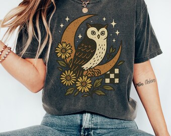 May include: Dark gray t-shirt featuring a whimsical owl perched on a crescent moon, surrounded by stars and sunflowers. The owl is brown and cream, the moon is tan, and the flowers are yellow. The shirt has a vintage, worn look.
