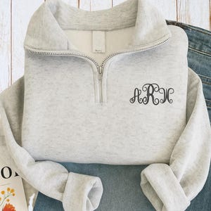 May include: A light gray quarter-zip sweatshirt with a silver zipper and a monogram in a script font. The sweatshirt is folded over a pair of blue jeans. The background is a light wood surface.