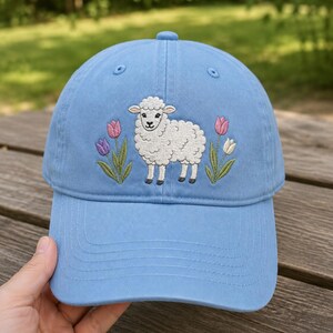 Könnte beinhalten: Eine hellblaue Baseballkappe mit einer Stickerei, die ein weißes Schaf in der Mitte zeigt, flankiert von zwei rosa Tulpen auf der linken Seite sowie einer rosa und einer weißen Tulpe auf der rechten Seite, mit grünen Stielen und Blättern.