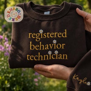 May include: A dark brown crewneck sweatshirt featuring yellow embroidered text that reads registered behavior technician. The design includes small white daisy accents between the words and on the sleeve, where the name Kayla is also embroidered.