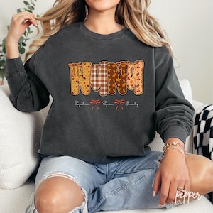 Op de afbeelding: Donkergrijze sweatshirt met het woord "NANA" in een herfstthema ontwerp. De letters zijn versierd met patronen van ruitjes, bladeren en patchwork. Onder het woord staan de namen "Sophia", "Ryan" en "Emily".