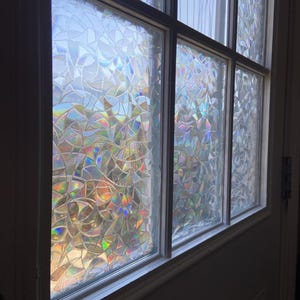 Op de afbeelding: Een raam met meerdere panelen met een decoratieve film. De film creëert een mozaïekachtig patroon met iriserende, regenboogkleurige reflecties. De raamkozijn is wit, wat een spel van licht en kleur creëert.