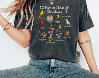 May include: Dark gray t-shirt featuring the text "12 Festive Birds of Christmas" with illustrations of various birds. The shirt has a crew neck and short sleeves. The design includes a canary, cardinal, hummingbird, and other birds.