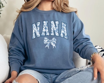 May include: A blue sweatshirt with a floral print design that says "NANA" in white letters with a white bow underneath.