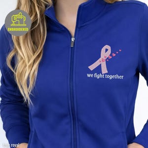 May include: True royal blue zip-up jacket with an embroidered pink ribbon and hearts design. The text "we fight together" is below the ribbon. The jacket has a high collar and a silver zipper.