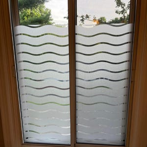 Peut inclure: Une fenêtre avec un film de confidentialité décoratif appliqué. Le film présente un motif de vagues répétées de couleur blanc givré, créant un effet texturé. La fenêtre est encadrée dans une couleur bois clair, avec une vue sur les arbres et le ciel visible à travers le verre.