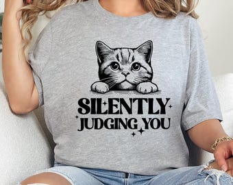Może przedstawiać: Szary T-shirt z czarno-białym obrazkiem kota patrzącego zza górnej części koszulki. Pod kotem nadrukowany jest tekst "SILENTLY JUDGING YOU".