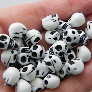 May include: A collection of small, white skull-shaped beads with black detailing. The beads have a glossy finish and are designed with eye sockets, a nose, and a detailed jawline. These beads are ideal for jewelry making or craft projects.