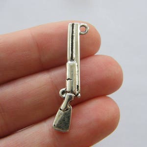 Könnte beinhalten: Silberfarbener Metallanhänger in Form einer Schrotflinte mit einer Öse oben zum Anbringen an einer Halskette oder einem Armband.