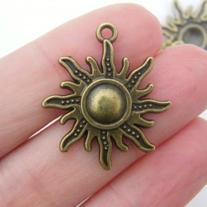 Könnte beinhalten: Antiker bronzefarbener Sonnenanhänger mit einer zentralen Scheibe und strahlenförmigen Strahlen. Das Sonnenstrahl-Design weist kleine Punktdetails entlang der Strahlen und eine Schlaufe zur Befestigung an Schmuck oder anderen Bastelarbeiten auf.