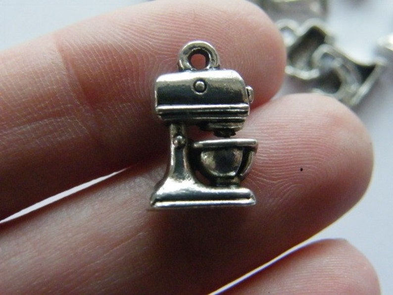 May include: Silver charm in the shape of a stand mixer. The charm is about 2 inches tall and has a loop at the top for attaching to a bracelet or necklace.