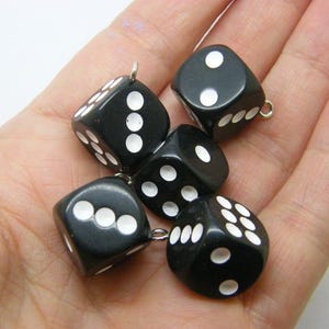 Puede incluir: Cinco dados negros con puntos blancos, cada uno con un pequeño lazo de metal adjunto. Los dados se muestran en una mano, mostrando varias combinaciones de puntos. Un accesorio de juego clásico.