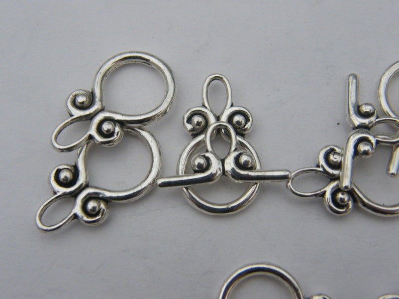 Pu&ograve; includere: Chiusure in metallo argentato con disegni decorativi e piccoli accenti rotondi. Queste chiusure sono utilizzate per la creazione di gioielli e l'artigianato.