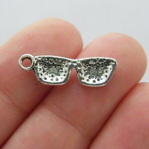 May include: Silver charm in the shape of sunglasses. The charm has a textured surface and a loop for attaching to a bracelet or necklace.