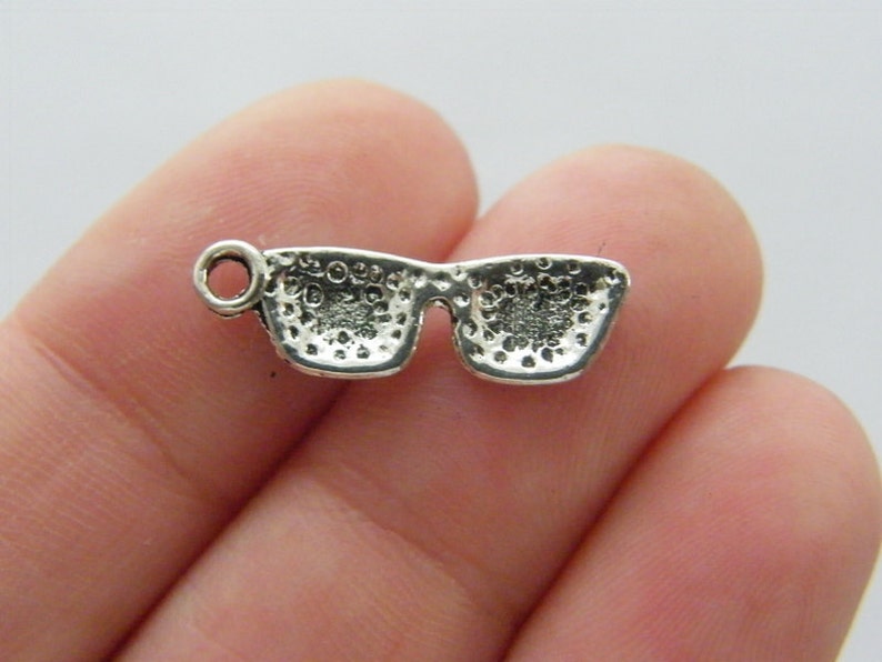May include: Silver charm in the shape of sunglasses. The charm has a textured surface and a loop for attaching to a bracelet or necklace.
