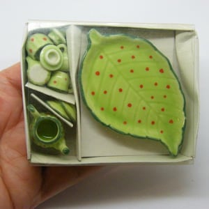 May include: A miniature tea set in a clear plastic box. The set includes a teapot, cups, saucers, and a leaf-shaped plate with red polka dots. The tea set is made of green and white ceramic.