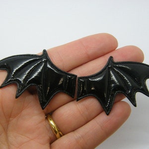 May include: A pair of glossy black bat wing appliques. Each wing features a pointed design with detailed veining and a shiny surface. These appliques are suitable for crafting or costume design, adding a gothic or Halloween theme.