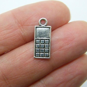 Puede incluir: Un pequeño dije plateado con forma de teléfono móvil vintage. El dije tiene un cuerpo rectangular con una pantalla en la parte superior y un teclado de botones cuadrados debajo. En la parte superior hay un pequeño lazo para sujetarlo a una cadena o pulsera.