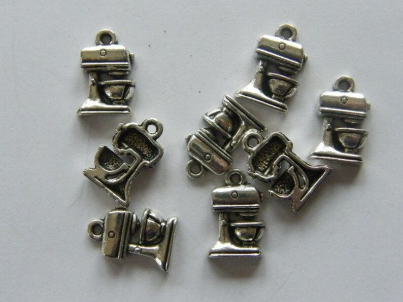 May include: A group of nine silver-toned metal charms shaped like a stand mixer. The charms have a detailed design and are perfect for adding a touch of kitchen-themed charm to jewelry or crafts.