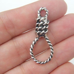 Puede incluir: Un colgante plateado con forma de soga corrediza. La sección superior es un nudo apretado, que se transforma en un diseño de cuerda trenzada que forma el lazo. El colgante es probablemente de metal.