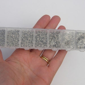 Puede incluir: Una caja de almacenamiento de plástico transparente con siete compartimentos llenos de anillos de salto plateados. La caja está en la mano de una persona.