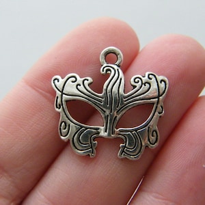 Puede incluir: Un dije de máscara plateada y ornamentada, con un lazo en la parte superior para colgar. La máscara presenta un diseño simétrico con patrones arremolinados y aberturas para los ojos, ideal para bisutería o manualidades.