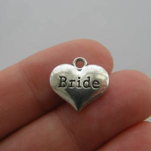 Puede incluir: Un charm plateado en forma de corazón con la palabra "Bride" grabada en el centro. El charm tiene un pequeño lazo en la parte superior para sujetarlo a un collar o pulsera. Un accesorio temático de boda.