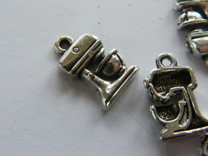 May include: Two silver metal charms shaped like a stand mixer. The charms have a detailed design and are perfect for adding a touch of kitchen-themed charm to jewelry or crafts.