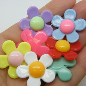 以下が含まれることがあります： 盛り上がった中心を持つカラフルなプラスチック製のフラワーシェイプボタンの詰め合わせ。ボタンは黄色、ピンク、赤、青、緑、紫の色合いです。