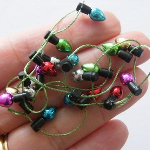 Puede incluir: Una cadena de luces navideñas en miniatura con bombillas de colores en rojo, verde, azul, rosa, dorado y plateado. Las luces están enhebradas en un cordón verde con conectores negros.