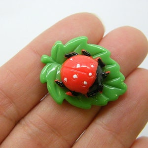 Può includere: Una coccinella rossa con macchie nere e zampe nere è seduta su una foglia verde. La coccinella e la foglia sono di plastica.
