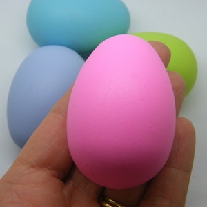 May include: A hand holding a bright pink egg, with other pastel-coloured eggs in the background. The eggs are in shades of blue and green. The eggs are smooth and solid, suggesting a decorative or craft item.